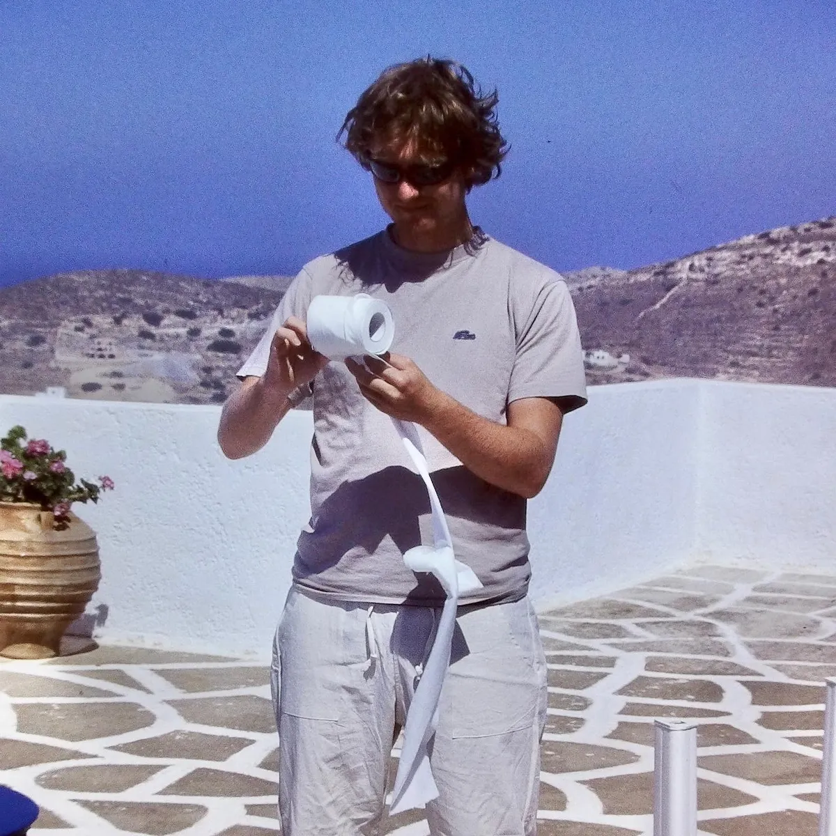 Man in white t-shirt holding toilet paper roll on white stone terrace with decorative pottery and hills in background, Ios, Greece