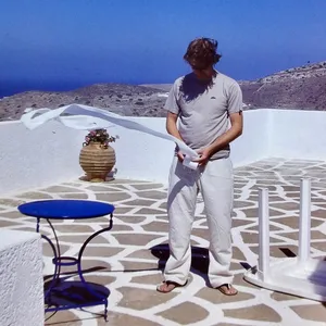 Person in grey shirt and white pants standing on white stone terrace with blue table and pottery, overlooking barren hillside and blue sea in Ios, Greece