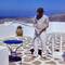 Person in grey shirt and white pants standing on white stone terrace with blue table and pottery, overlooking barren hillside and blue sea in Ios, Greece