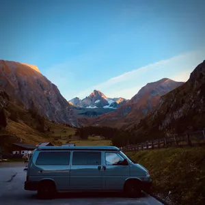 Blue VW T4 van parked on mountain road with snow-capped Austrian Alps and Großglockner peak in background during golden hour