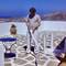Person in grey t-shirt and white pants holding white mop on geometric patterned stone terrace with pottery and blue table, Ios Greece