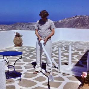 Person in grey t-shirt and white pants holding white mop on geometric patterned stone terrace with pottery and blue table, Ios Greece
