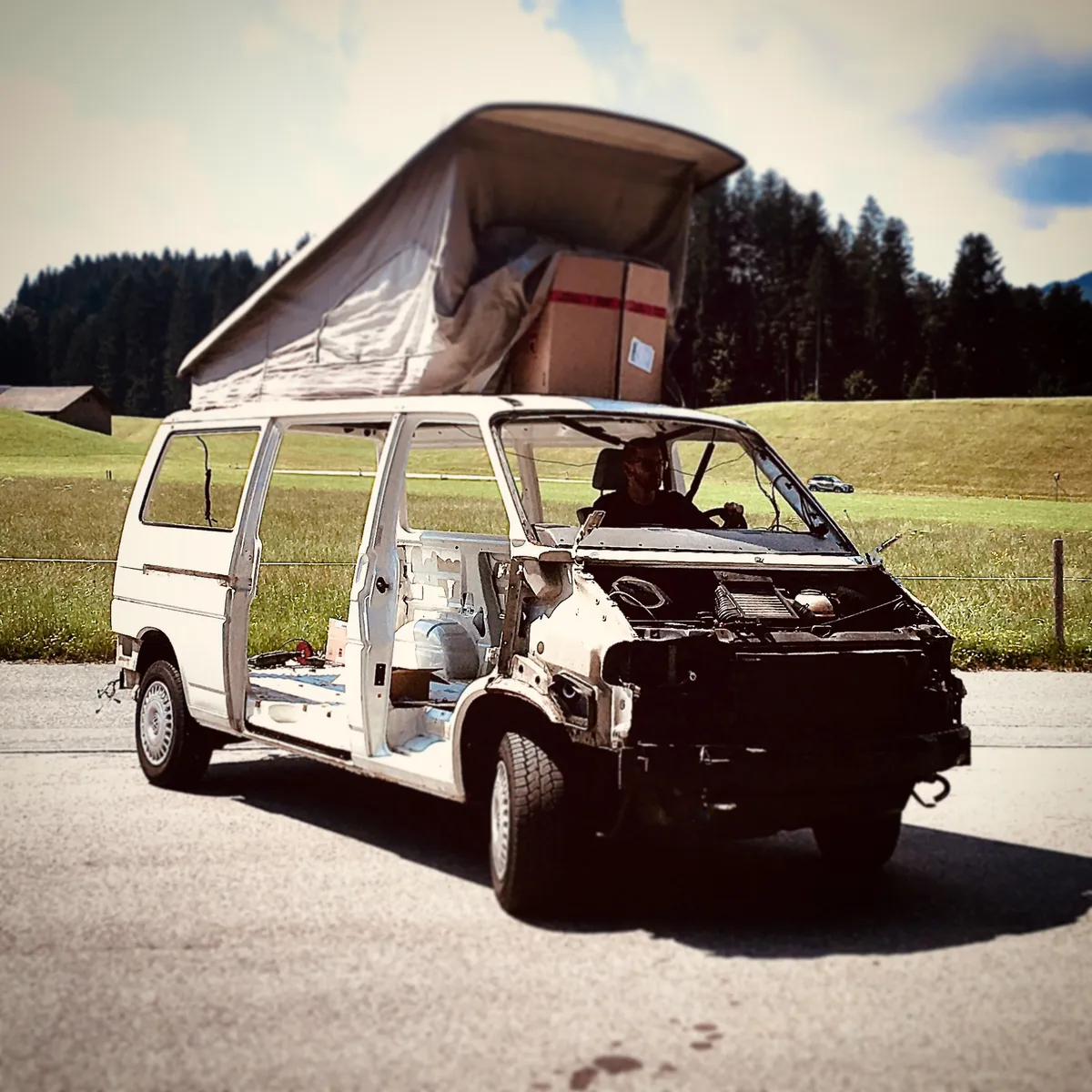 White Volkswagen T4 van with front end removed and pop-up tent on roof, parked on road with green fields and forest in background, Kitzbühel Austria