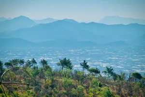 Panoramic view of multiple mountain ranges receding into hazy blue distance, with green forest in foreground and valley settlements below, taken from elevated viewpoint in Italy