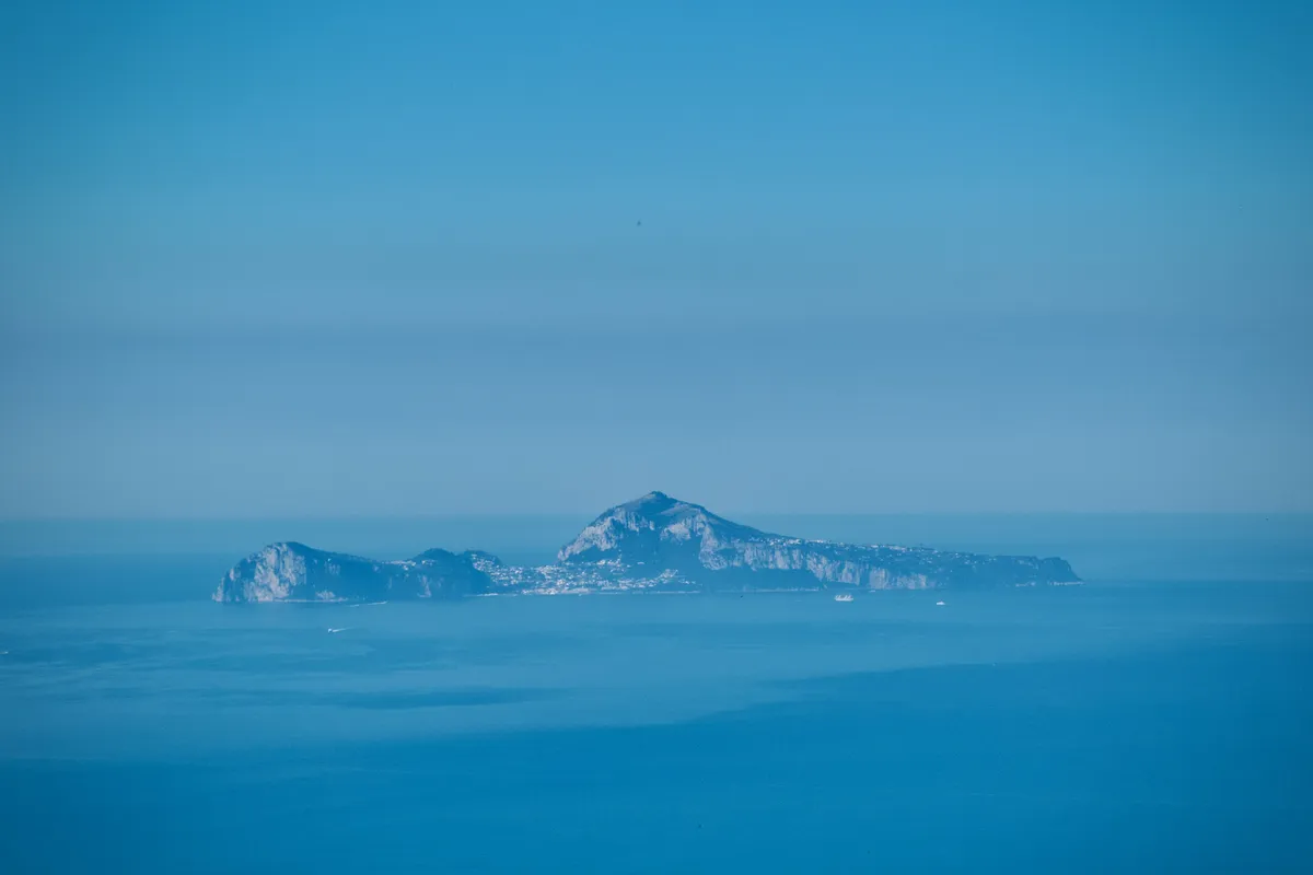 Distant view of Capri island silhouetted against blue sky across Gulf of Naples waters, photographed from Mount Vesuvius, Italy