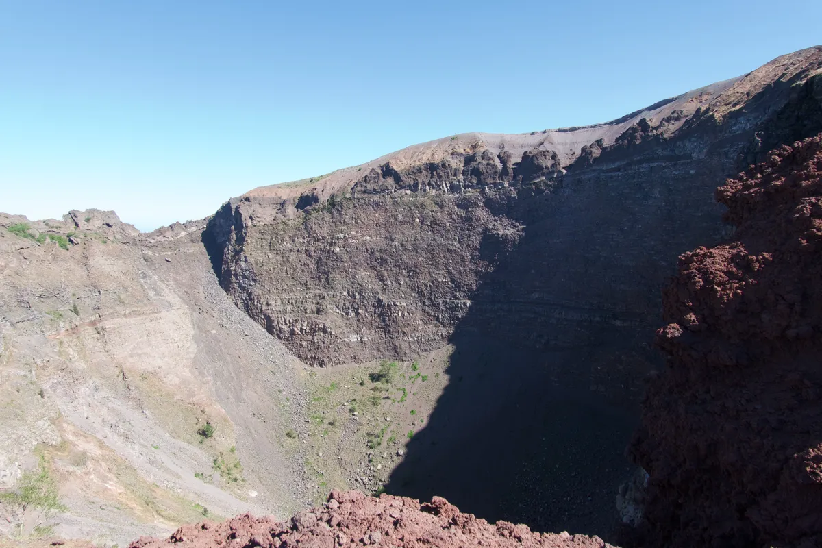 Deep volcanic crater of Mount Vesuvius showing steep rocky walls with visible geological layers and sparse green vegetation on crater floor, Naples, Italy