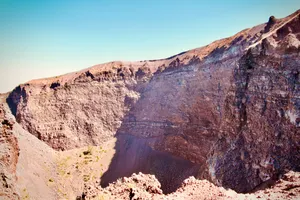 View down into Mount Vesuvius crater showing steep stratified rock walls, dark crater floor, and small patches of vegetation, Ottaviano, Italy