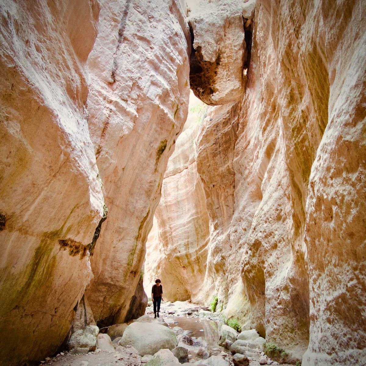 Person walking through narrow slot canyon with towering cream and rust-colored sandstone walls, scattered rocks on canyon floor, Pano Arodes, Cyprus