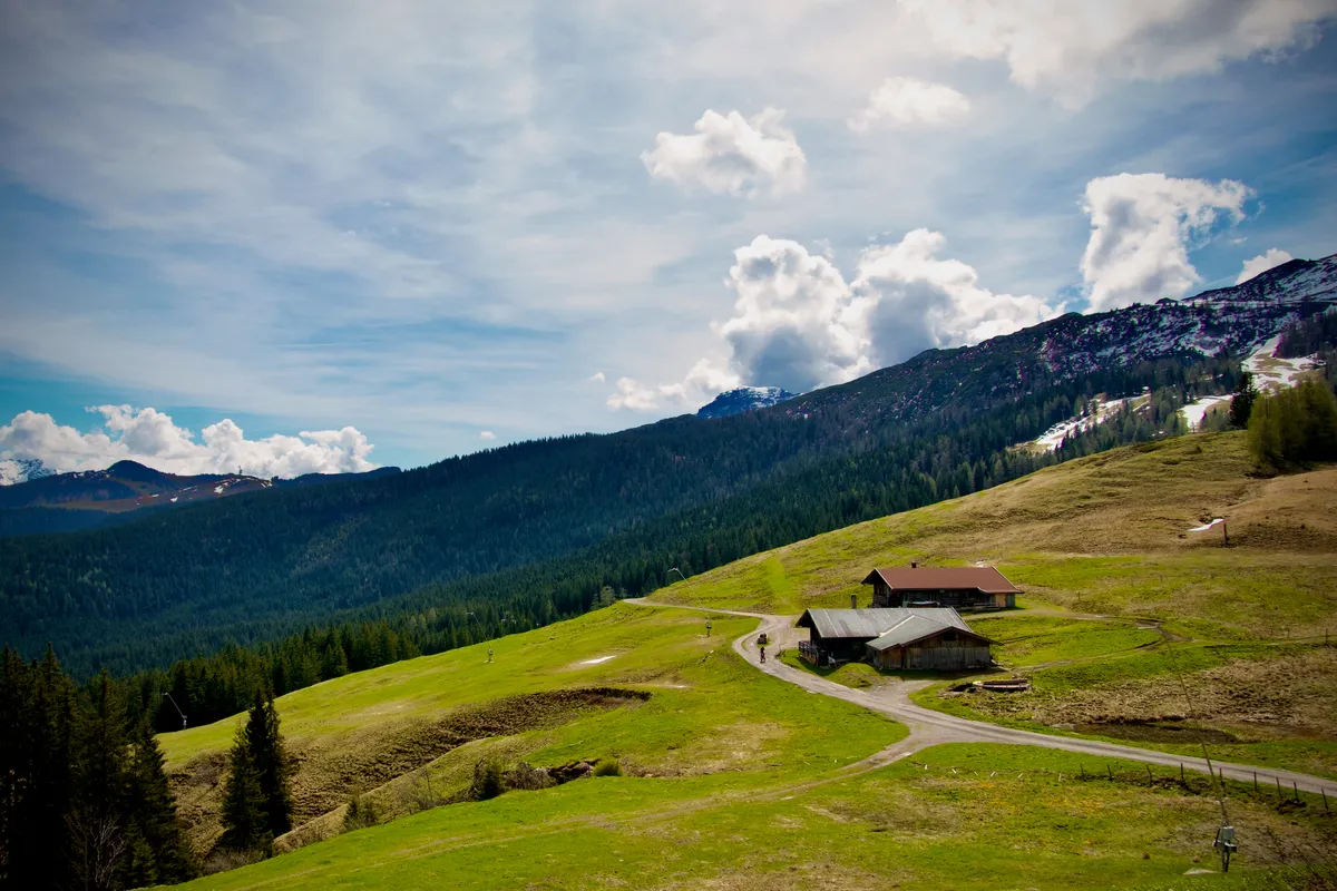 Alpine meadow landscape with traditional wooden buildings, winding dirt road, forested mountains and snow-capped peaks under cloudy sky near Steinplatte, Austria