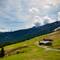Alpine meadow landscape with traditional wooden buildings, winding dirt road, forested mountains and snow-capped peaks under cloudy sky near Steinplatte, Austria