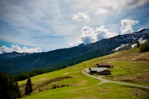 Alpine meadow landscape with traditional wooden buildings, winding dirt road, forested mountains and snow-capped peaks under cloudy sky near Steinplatte, Austria