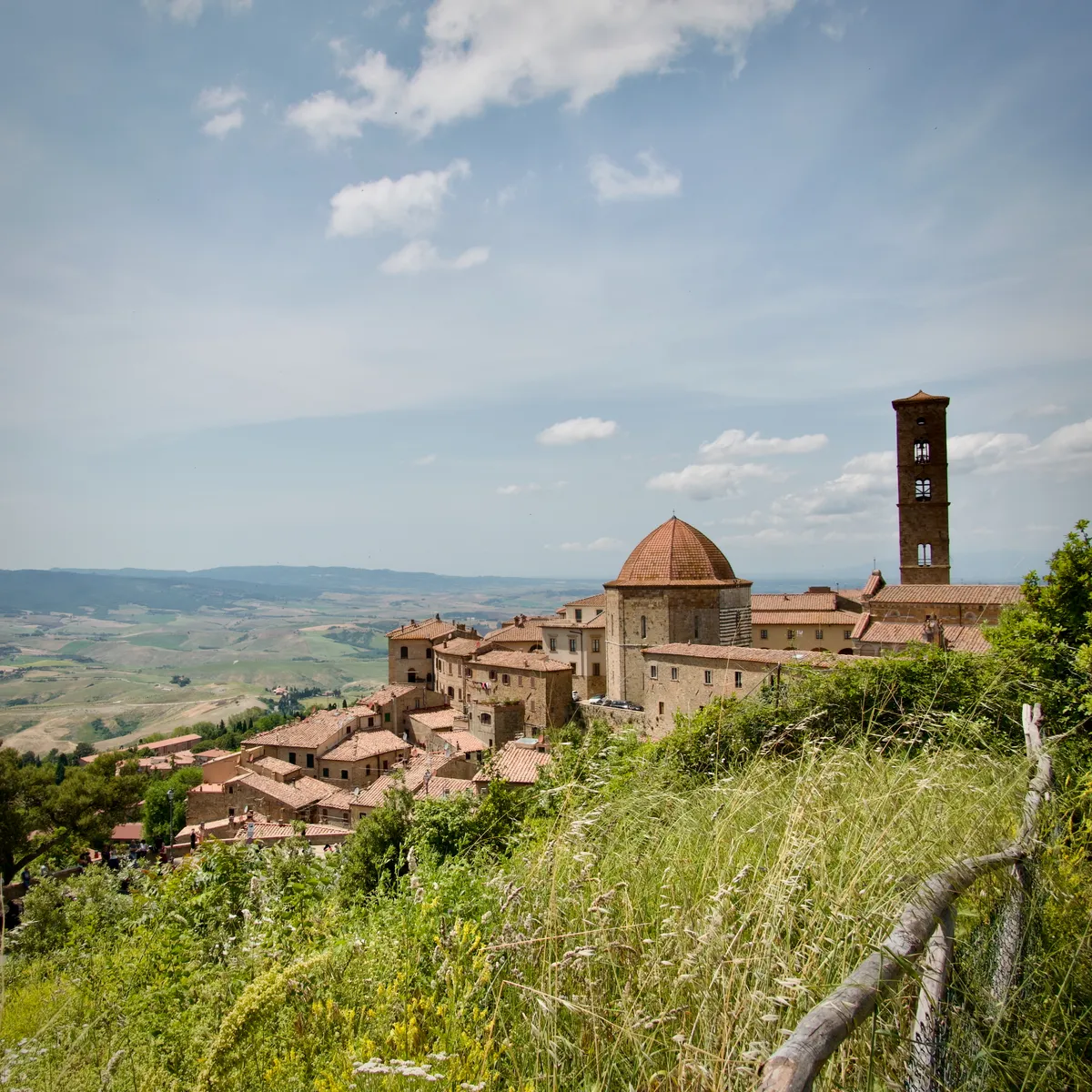 Medieval bell tower and domed church rising above clustered stone buildings in hilltop town of Volterra, Italy, viewed through wild grass and wooden fence with countryside beyond