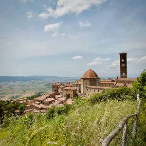 Medieval bell tower and domed church rising above clustered stone buildings in hilltop town of Volterra, Italy, viewed through wild grass and wooden fence with countryside beyond