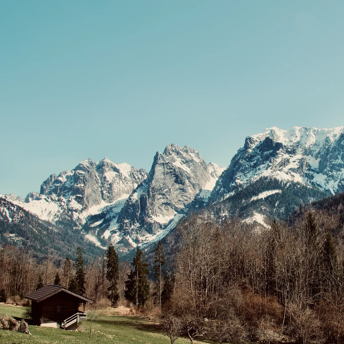 Snow-covered Wilder Kaiser mountain peaks rising dramatically above valley floor with traditional wooden hut and bare trees in Kaisertal, Austria