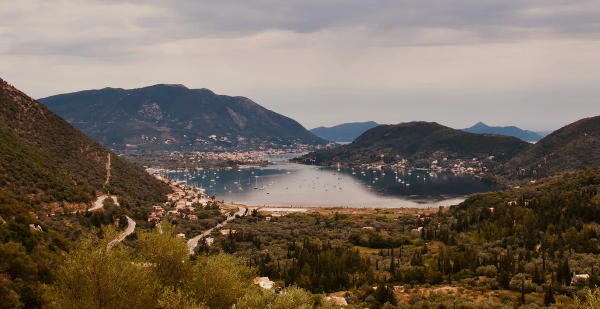 Aerial view of a Mediterranean harbor in Lefkada, Greece, surrounded by green hills with scattered white buildings and numerous boats anchored in calm blue water