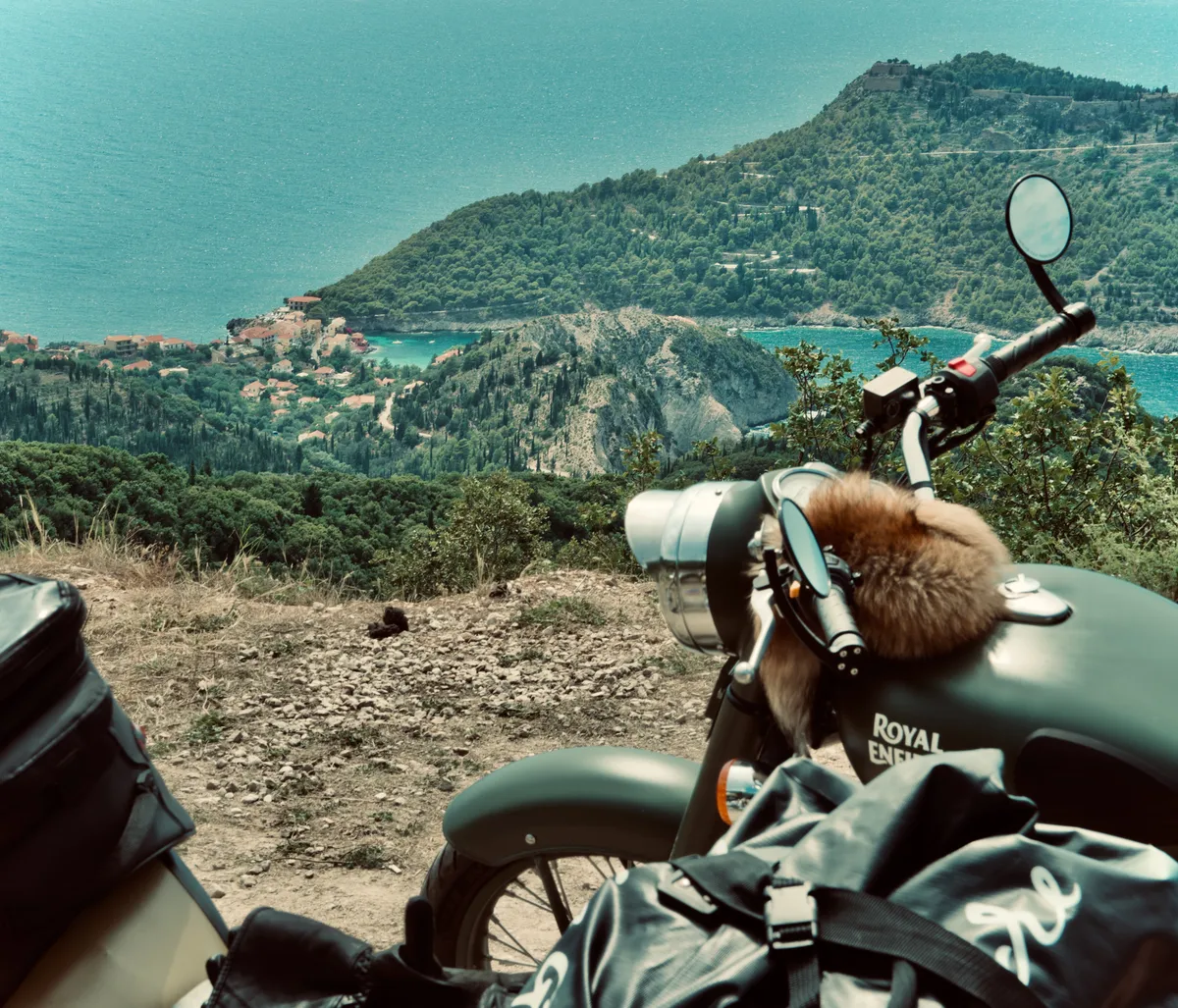 Royal Enfield motorcycle with furry animal on seat overlooking Assos coastline and turquoise Ionian Sea in Kefalonia, Greece