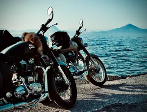 Two Royal Enfield motorcycles parked on coastal road overlooking blue water and mountains in Rio, Greece. One bike has fur accessory on seat.