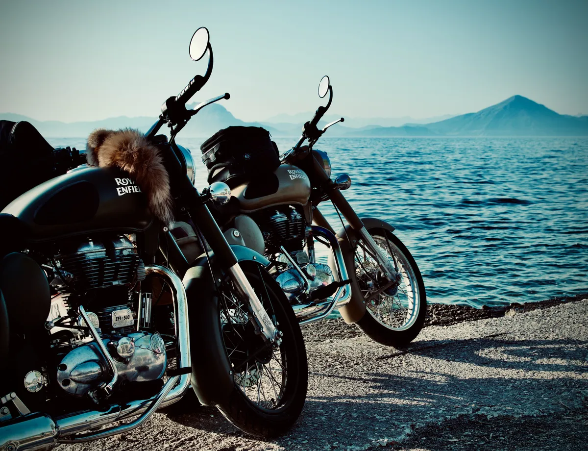 Two Royal Enfield motorcycles parked on coastal road overlooking blue water and mountains in Rio, Greece. One bike has fur accessory on seat.