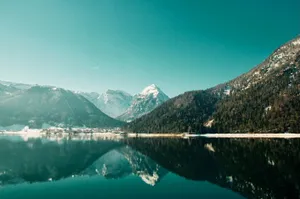 Pristine alpine lake reflecting snow-covered mountains and forested slopes under clear turquoise sky in Achensee, Austria