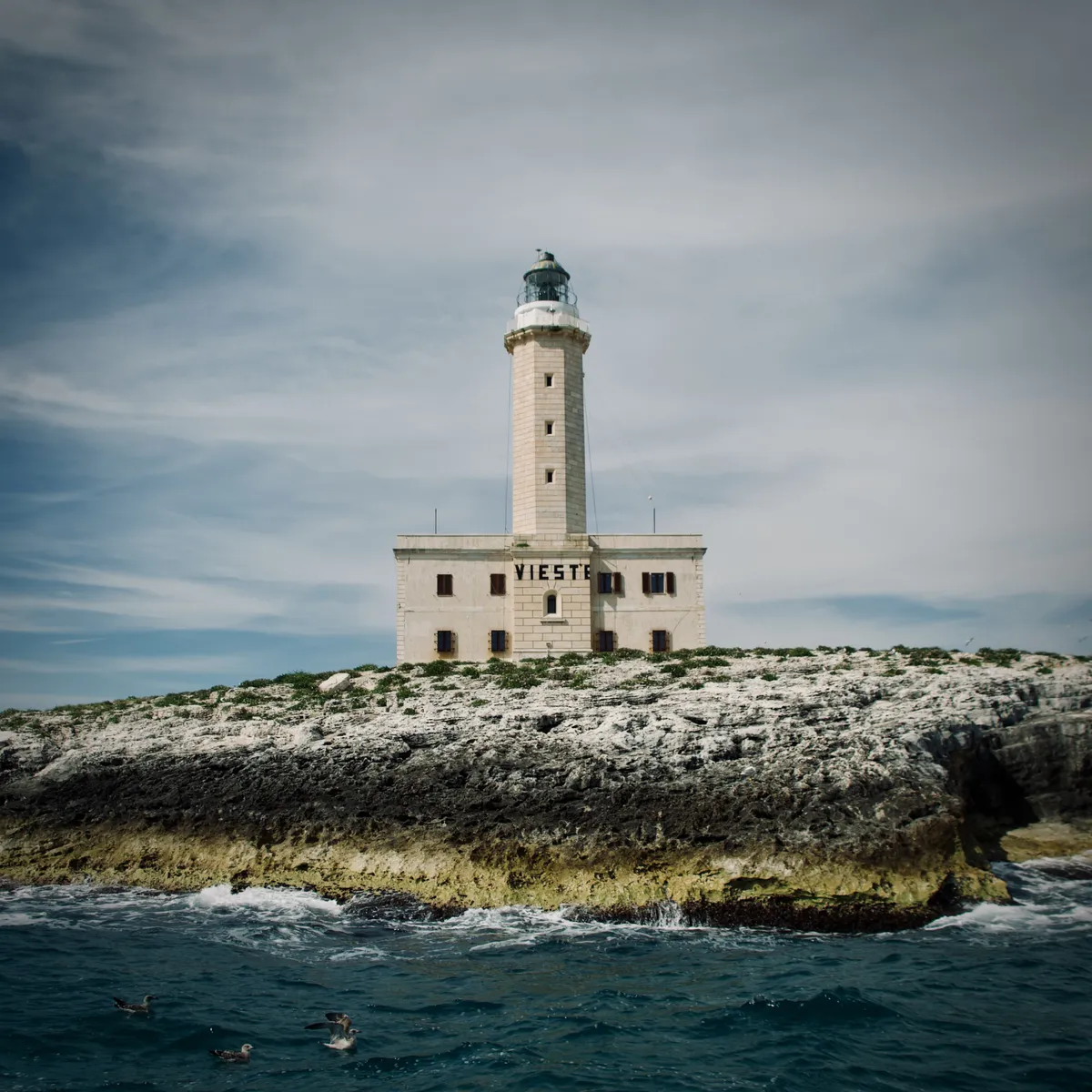 White limestone lighthouse with attached building on rocky coastal cliffs in Vieste, Italy, surrounded by blue-green Mediterranean waters