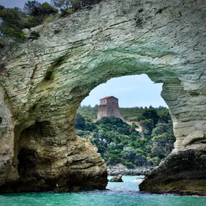 View through natural stone arch showing medieval tower on cliff overlooking turquoise Adriatic Sea in Vieste, Italy