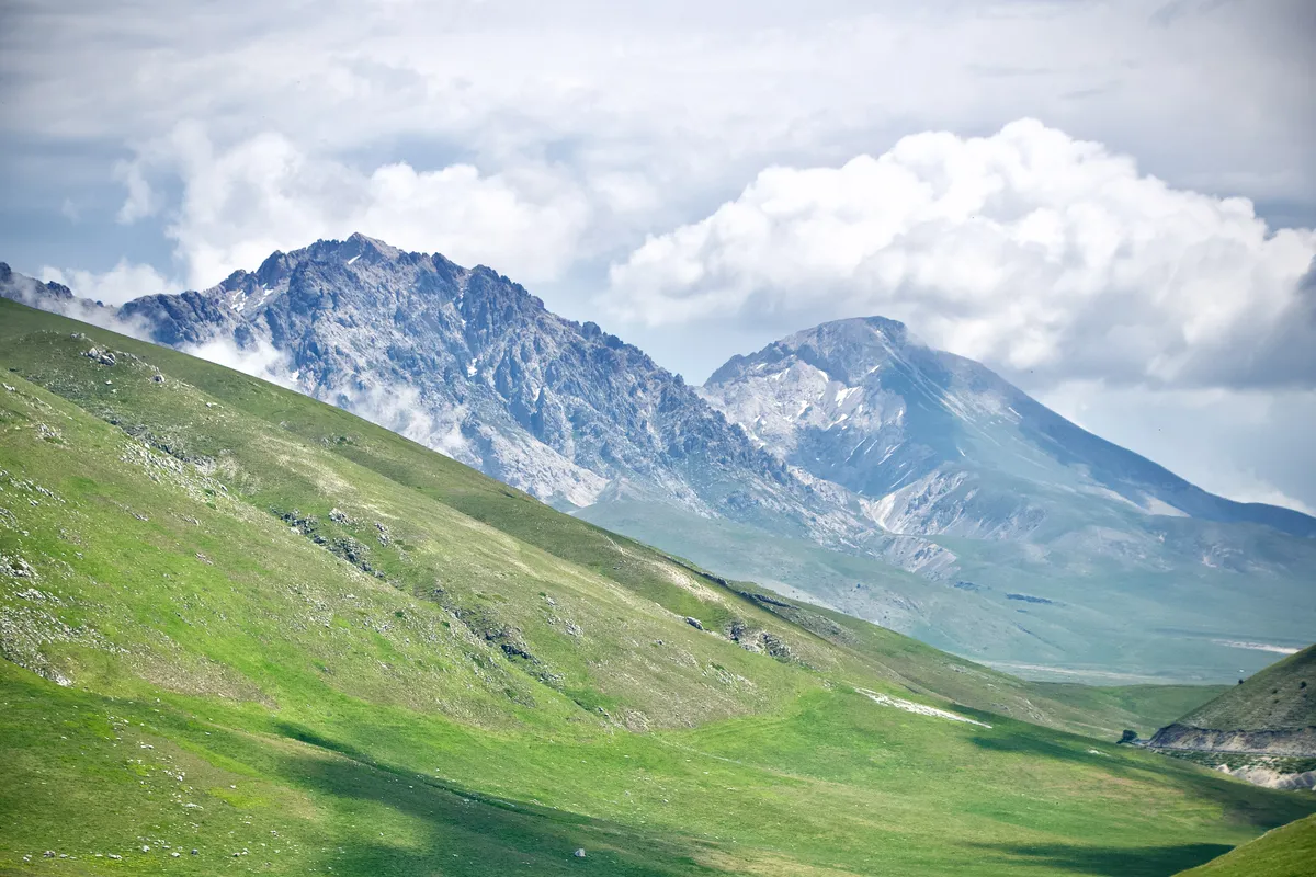 Rolling green hills leading to dramatic limestone mountain peaks under cloudy sky in L'Aquila, Abruzzo, Italy