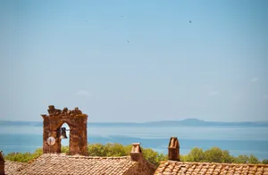 Stone bell tower with bell visible through arched opening, terracotta tile rooftops below, Lake Bolsena and hills in background, Bolsena, Italy