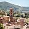 Tall stone bell tower with arched openings rising above clustered medieval buildings in Bolsena, Italy, with forested hills in background