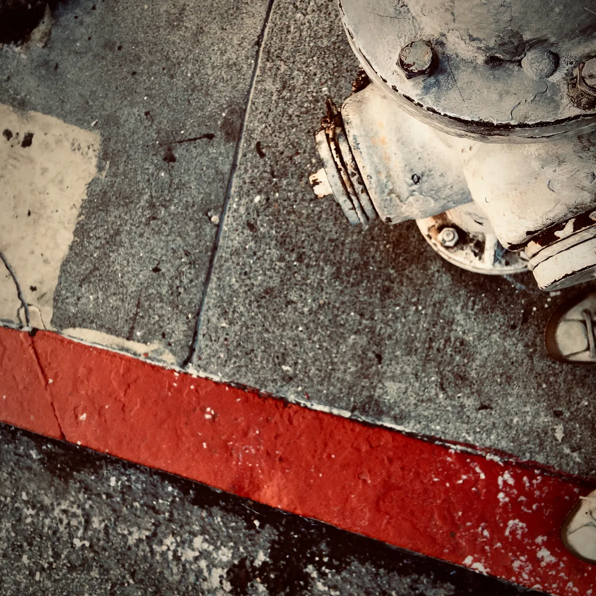 Red painted curb stripe on concrete sidewalk with weathered circular metal utility fixture and rusty bolts