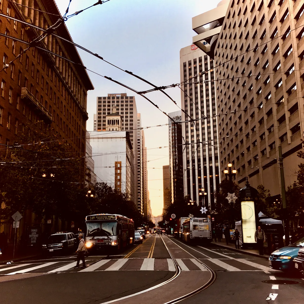 Downtown San Francisco street view with cable car overhead wires, tall buildings on both sides, buses and traffic during evening rush hour