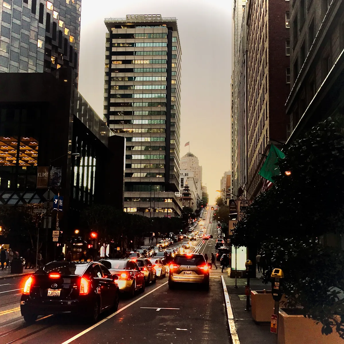 Heavy evening traffic with red brake lights on steep San Francisco street, tall buildings on both sides, cars backed up in multiple lanes