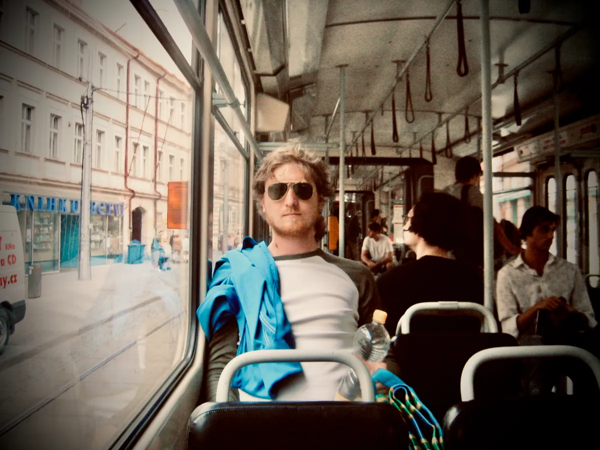Man in sunglasses and white shirt sitting on Prague tram interior, holding blue jacket, with European buildings visible through windows