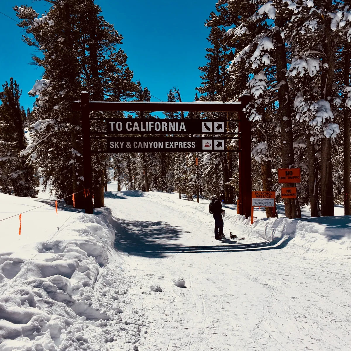 Wooden sign reading 'TO CALIFORNIA' and 'SKY & CANYON EXPRESS' on snowy mountain road with snow-covered pine trees and person with snowshoes in Kingsbury, California