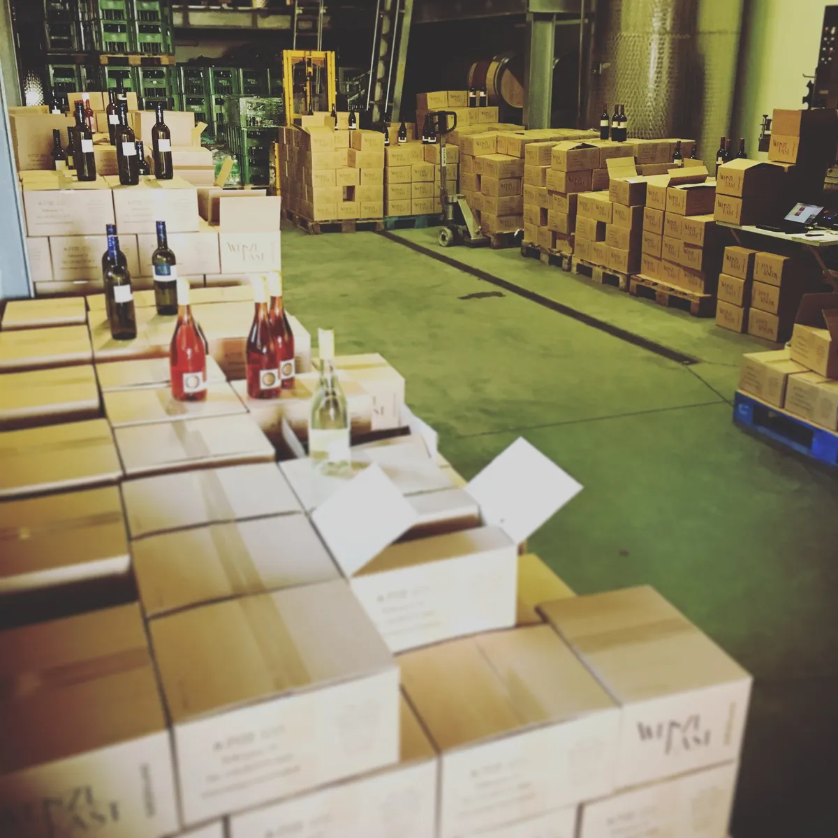 Wine storage warehouse with stacked cardboard boxes of wine bottles, sample bottles on floor, industrial shelving in Gols winery cellar