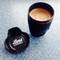 Black camera lens-shaped coffee mug filled with frothy coffee, with removable lid beside it on speckled blue surface