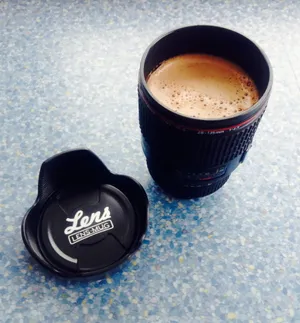Black camera lens-shaped coffee mug filled with frothy coffee, with removable lid beside it on speckled blue surface