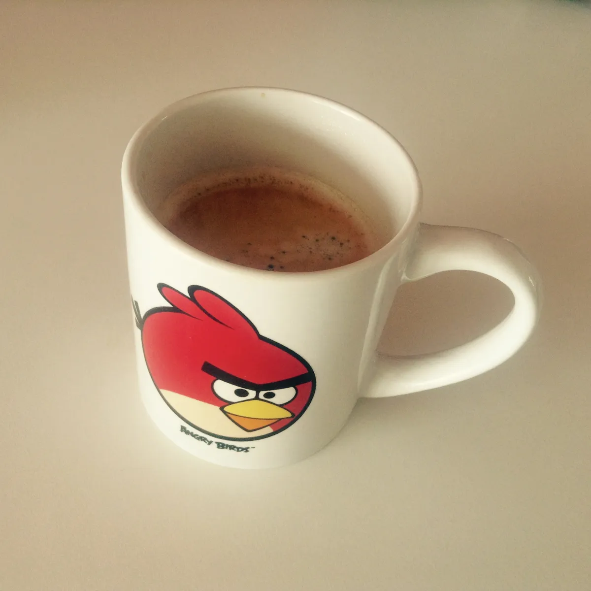 Small white coffee mug with red Angry Birds character design containing brown coffee with foam, photographed on light colored surface