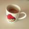 Small white coffee mug with red Angry Birds character design containing brown coffee with foam, photographed on light colored surface