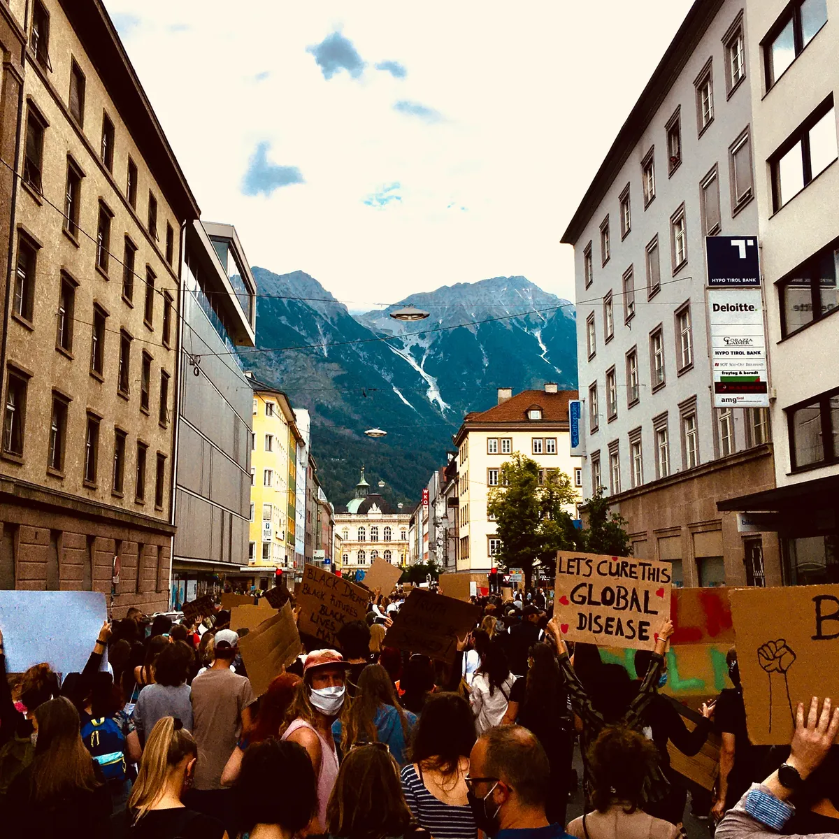 Black Lives Matter protest in Innsbruck, Austria with crowds holding cardboard signs including 'Let's Cure This Global Disease' against Alpine mountain backdrop