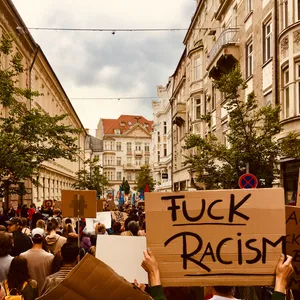 Anti-racism protest in historic Innsbruck city center with prominent cardboard sign reading 'FUCK RACISM' held above crowd of demonstrators