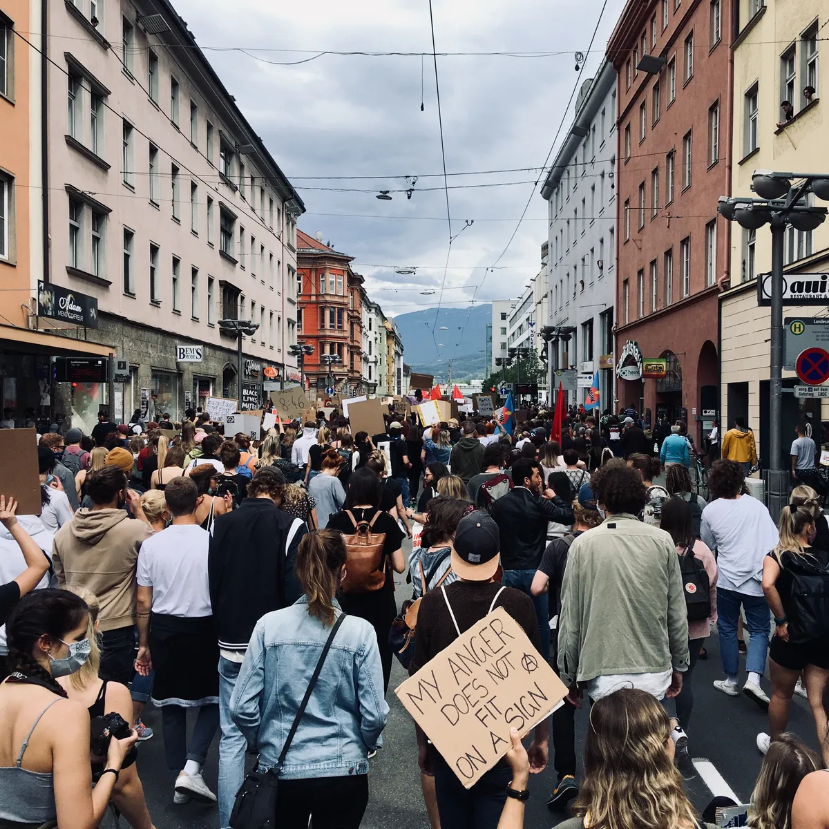 Large crowd of Black Lives Matter protesters filling pedestrian street in Innsbruck city center, carrying cardboard signs including 'MY ANGER DOES NOT FIT ON A SIGN'