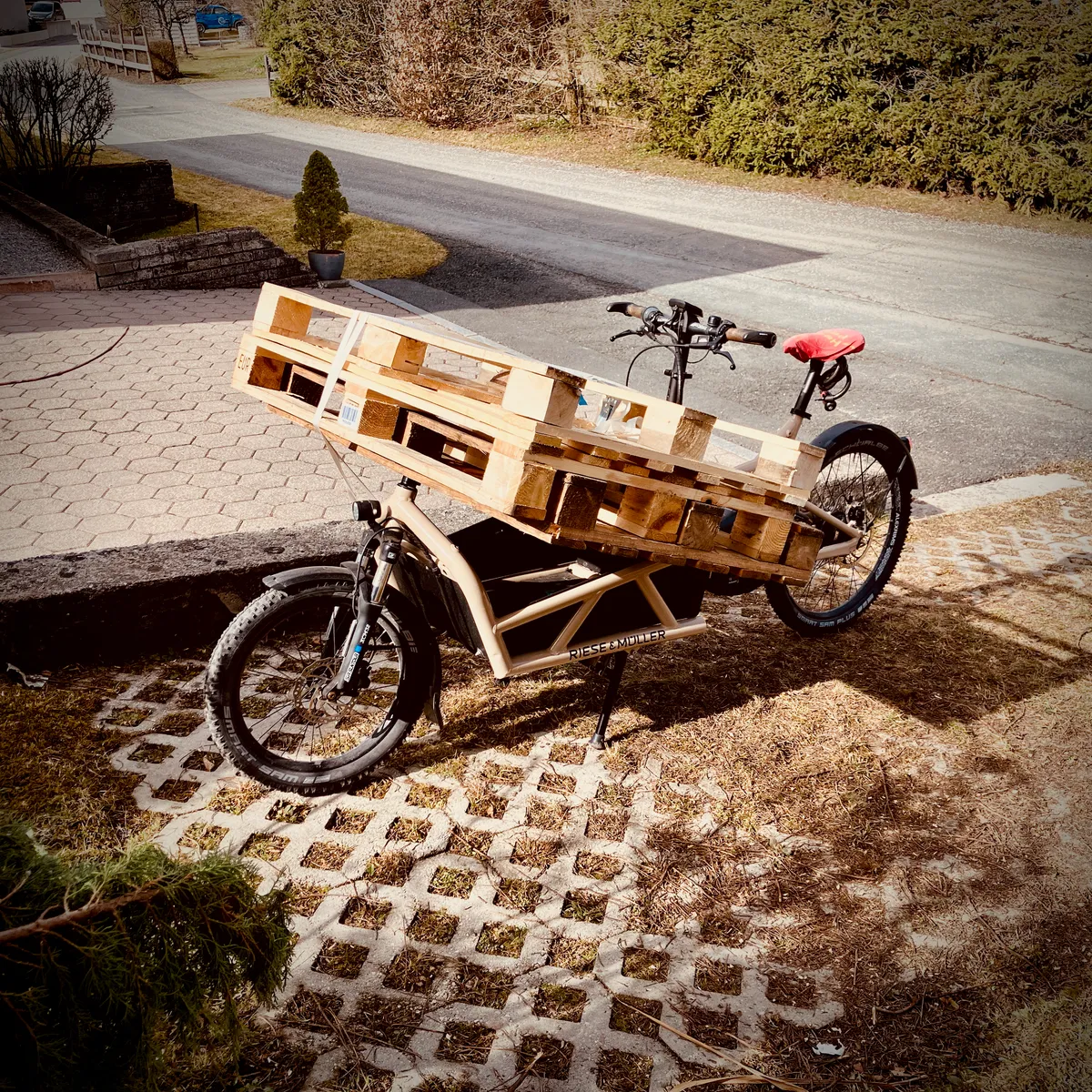 Riese & Müller cargo bike loaded with wooden pallets on wooden platform, parked on cobblestone driveway in residential area