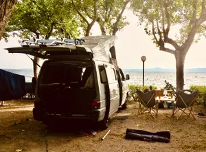 White VW T4 van parked under trees by Lake Bolsena, Italy, with camping chairs and gear set up nearby on sandy ground