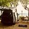 White VW T4 van parked under trees by Lake Bolsena, Italy, with camping chairs and gear set up nearby on sandy ground