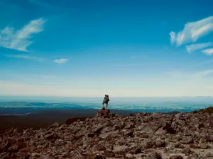Hiker with backpack standing on rocky summit outcrop overlooking vast landscape with rolling hills and blue sky, Ruapehu District, New Zealand