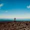 Hiker with backpack standing on rocky summit outcrop overlooking vast landscape with rolling hills and blue sky, Ruapehu District, New Zealand