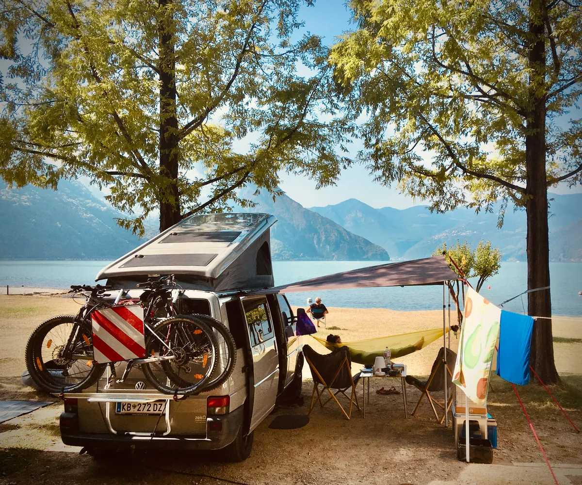 VW T4 camper van with pop-up roof and awning setup under trees at Lake Iseo, Italy, with mountains and lake in background