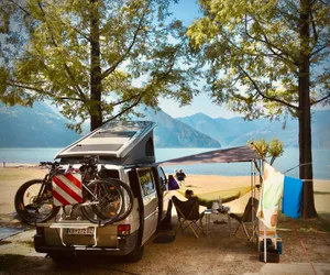 VW T4 camper van with pop-up roof and awning setup under trees at Lake Iseo, Italy, with mountains and lake in background