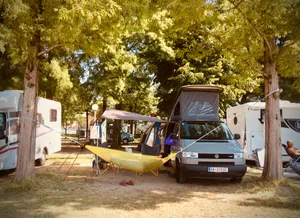 Yellow hammock stretched between trees next to white VW T4 camper van at campground in Pieve, Italy with other RVs visible