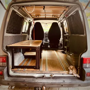 Interior view of 1996 VW T4 campervan showing wooden pull-out camping bed installation, with driver seats visible and open rear doors in Erlau, Austria